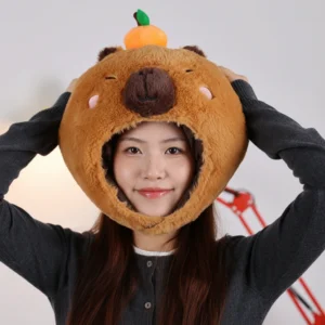 Model wearing a fluffy brown capybara plush hat with a small plush orange on top, showcasing the soft faux fur material and cute embroidered face.