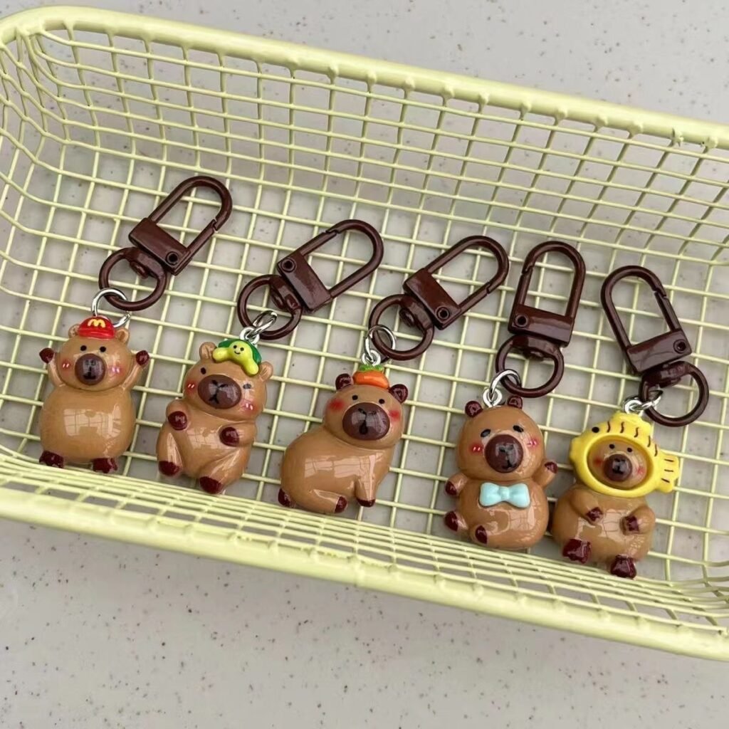 A set of five glossy resin capybara bag charms arranged in a row, featuring unique accessories including a fish hood, persimmon, green hat, blue bow tie, and red baseball cap.
