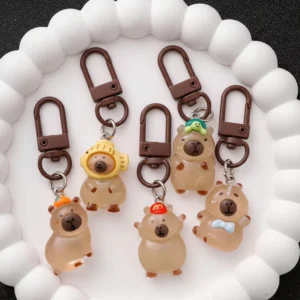 Set of 5 resin capybara bag charms shown in both daylight as translucent brown figures and in the dark glowing bright green, featuring designs with turtles, oranges, and hats.