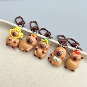 A set of five glossy resin capybara bag charms arranged in a row, featuring unique accessories including a fish hood, persimmon, green hat, blue bow tie, and red baseball cap.