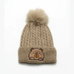 Beige cable knit winter hat for kids featuring a large fluffy pom-pom and a fuzzy embroidered capybara patch on the cuff.