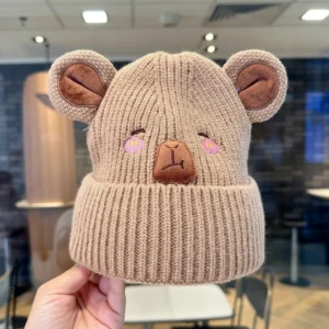 A camel baby knitted capybara beanie with large ears and embroidered pink blushing cheeks.