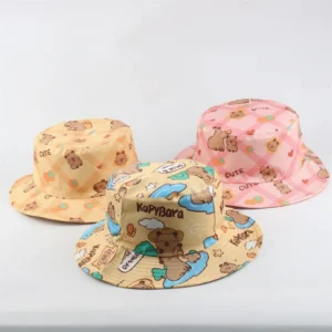 Three adult fabric bucket hats arranged together: one yellow plaid with capybaras, one pink plaid with stars, and one beige hat featuring a surfing capybara cartoon scene.