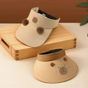 Two styles of beige adult hats: an open-top sun visor with 3D brown pom-poms on the brim, and a matching baseball cap with capybara ears, both featuring a cute face design.
