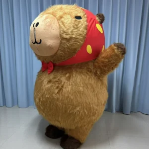 A full-body view of a brown plush inflatable capybara costume wearing a red polka dot headscarf and bow tie, waving its paw against a blue background.