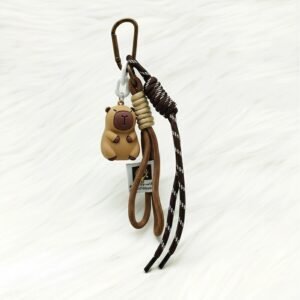 Close-up of a 3D PVC capybara keychain attached to a brown leather bag, featuring a utility rope strap and metal carabiner clip.