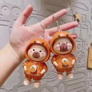Hand pressing the chest button on a cute PVC capybara keychain wearing an orange hoodie, demonstrating the face-changing mechanism.