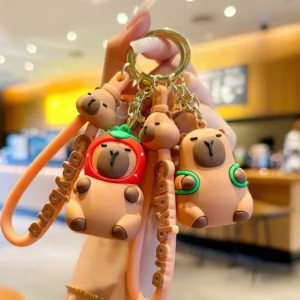 Two cute capybara keychains made of soft PVC, one wearing a red strawberry hood and the other wearing a green turtle backpack, featuring gold alloy hardware and matching straps.