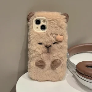 Capybara Plush Phone Case with Hair Clip A beige, long-pile fluffy phone case designed to look like a sleeping capybara with embroidered closed eyes, small brown paws, knitted ears, and a small decorative plastic hair clip attached to the fur.
