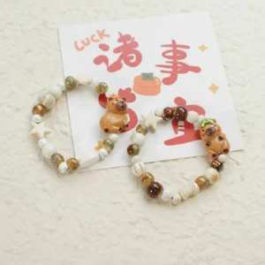 Two styles of elastic beaded bracelets featuring ceramic capybara charms, one with a persimmon on its head and one with a green frog, strung with star and bone-shaped beads in creamy earth tones.