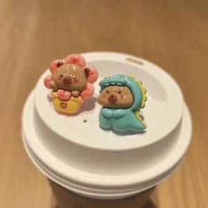 A pair of cute capybara stud earrings featuring one capybara in a yellow bee costume and another set with a pink flower and blue dinosaur design.