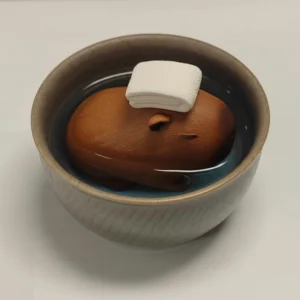 A brown textured resin capybara mini figure with a white towel on its head, sitting inside a ceramic tea bowl filled with water as a traditional tea pet.