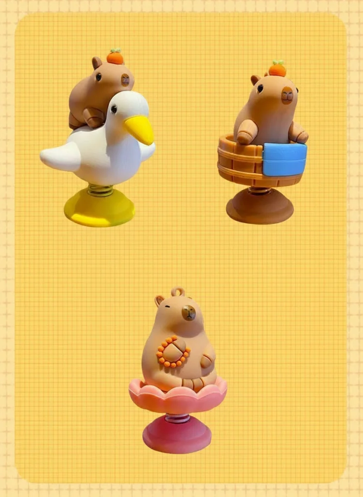 Four capybara bobblehead figures with different toppers—a bird, a sprout, an orange, and a flower—shown with their dimensions and silver metal spring bases.
