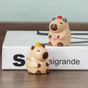 Two brown ceramic capybara mini figures in a meditative pose, one wearing a pink crown and the other holding prayer beads, displayed on a wooden surface.