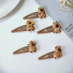 Flat lay of five brown metal snap hair clips featuring 3D resin capybara charms with a turtle, fish hat, red cap, bow tie, and mandarin orange.