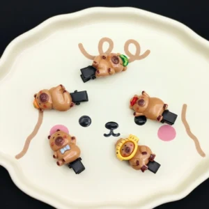 A set of five mini black plastic hair clips featuring small 3D resin capybara charms with various accessories like a turtle, fish hat, and red cap.