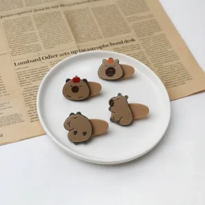 Top-down view of four flat acrylic capybara hair clips: two featuring front faces with fruit on top, and two showing side-profile body poses.