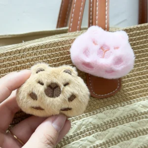 Close-up of a soft, fuzzy capybara head brooch in brown and pink colors, featuring embroidered facial details and a plush texture.