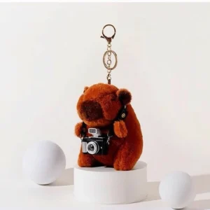 Front view of a brown capybara plush toy wearing a black camera around its neck with a gold keychain attachment.