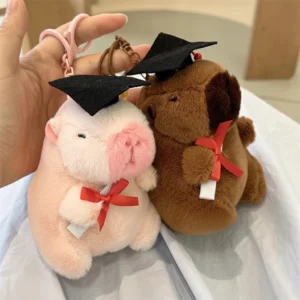 A brown and a pink capybara plush keychain sitting side by side, both wearing black graduation caps and holding diplomas with red ribbons.
