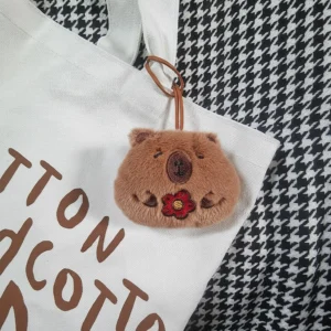 Capybara plush name tag hanging from a white canvas bag handle via a brown leather-style strap.