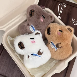 Three capybara plush keychains with different fur colors, each holding a small fish toy.
