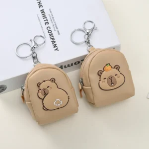 A beige faux leather capybara purse shaped like a mini backpack, shown in two styles: one with an orange on its head and one looking back, featuring silver keychain hardware.