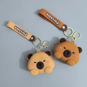 Silicone capybara-shaped earbud case and coin purse in tan and brown, featuring a wrist strap and shown holding white wireless earphones.