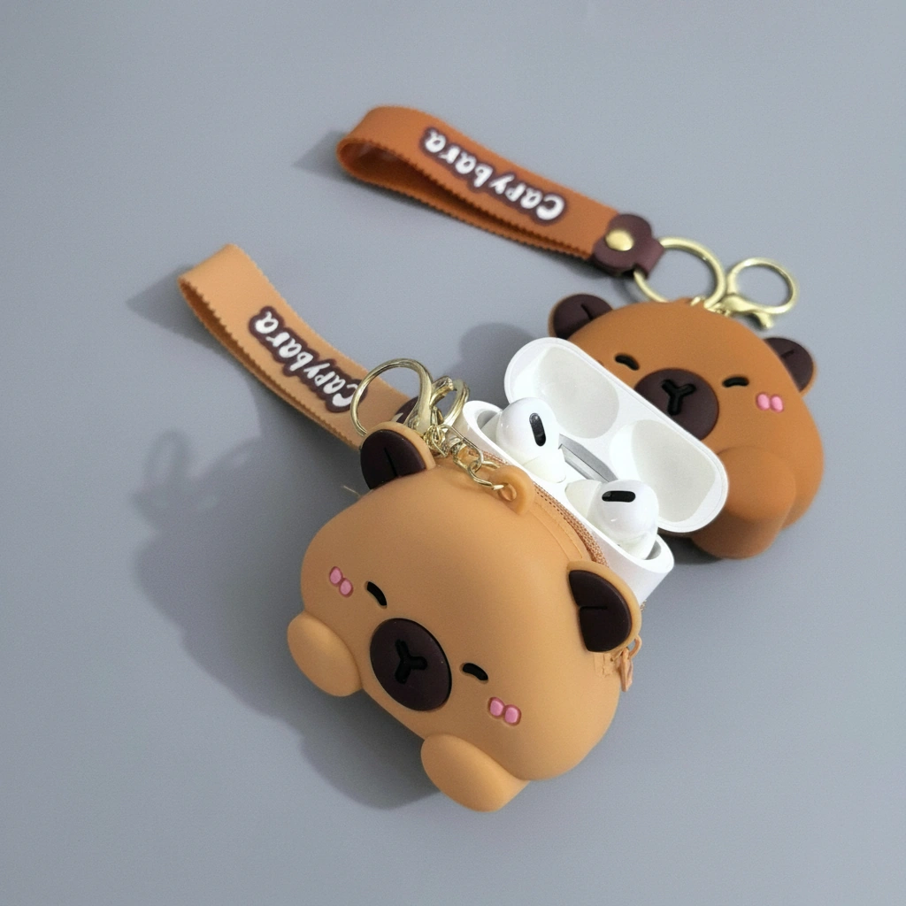 Silicone capybara-shaped earbud case and coin purse in tan and brown, featuring a wrist strap and shown holding white wireless earphones.